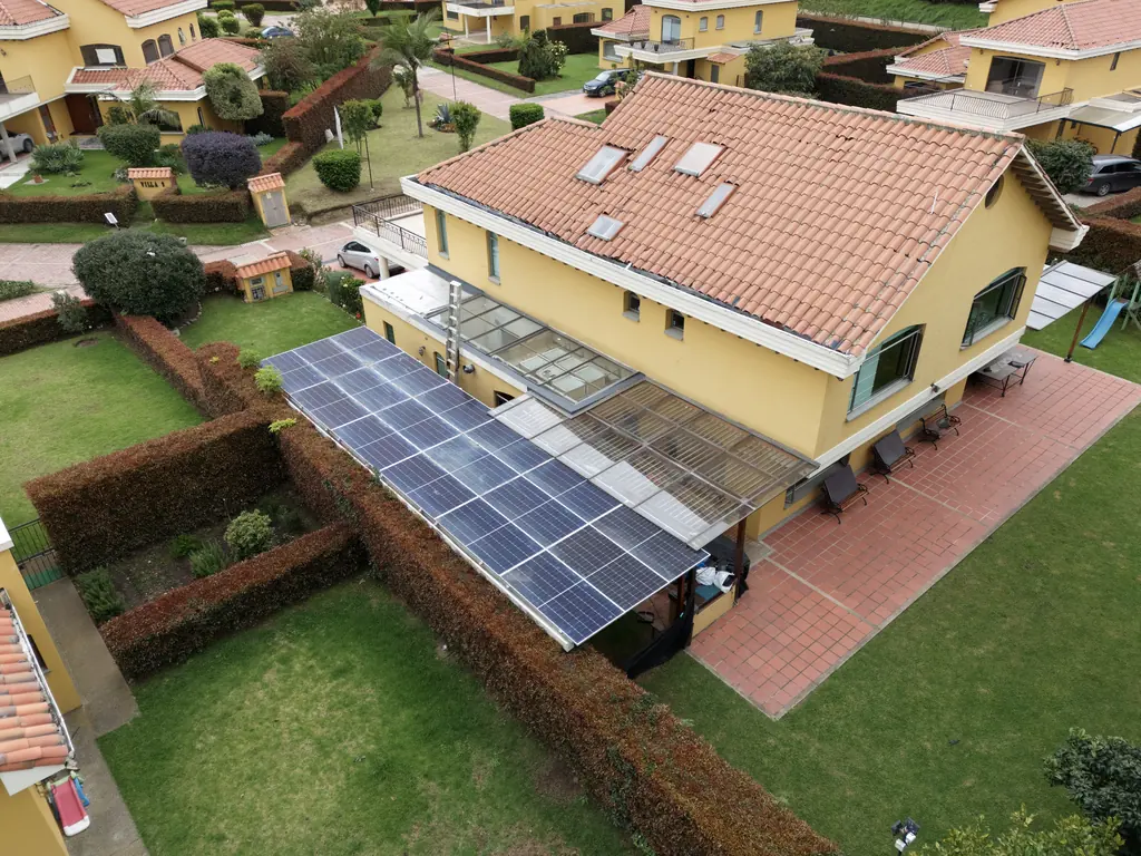 Vista de dron de la casa 15 en Pueblo Viejo, Cota: casa de techo de teja con una pérgola cubierta por dos filas de módulos solares junto al jardín.