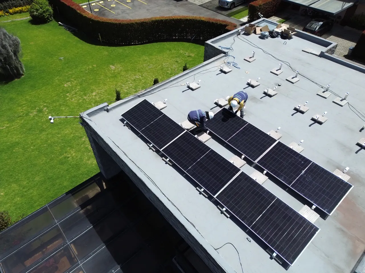 Vista de dron de un techo plano en un conjunto residencial de Cota: dos técnicos de SunWork fijan filas de módulos solares sobre estructura con lastre.
